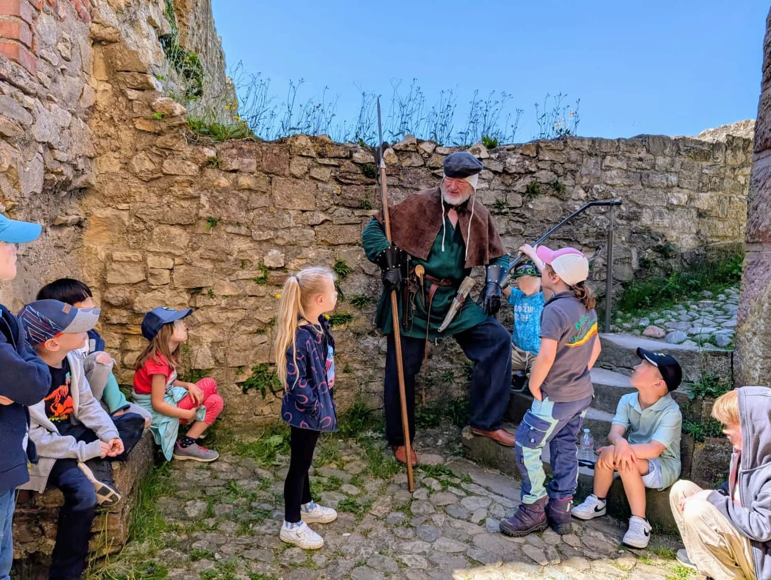 Der Burgknecht steht mit einigen Kindern unterhalb des Bergfrieds und erzählt ihnen von Geheimgängen auf der Burg.